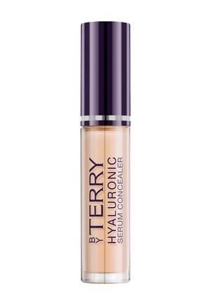 Tube transparent du correcteur By Terry Hyaluronic Serum avec un capuchon violet foncé et un liquide beige visible à l'intérieur sur fond blanc.