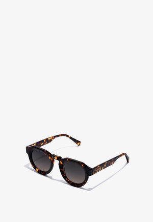 Okulary przeciwsłoneczne w kolorze musztardowym z okrągłymi soczewkami, ciemnym gradientowym przyciemnieniem, płaskim kształtem ramki i kontrastującymi jasnymi akcentami na zausznikach.