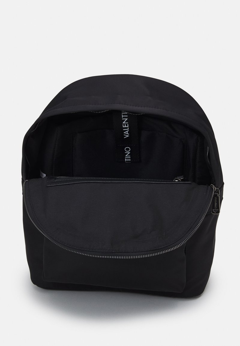 Mochila de tela negra con un diseño redondeado, que cuenta con una cremallera superior y un bolsillo interior. Incluye una correa de marca en su interior. Textura suave.