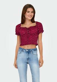 Junge Frau, die ein kastanienbraunes, kurzärmliges Polka-Dot-Crop-Top und helle, hochtaillierte Jeans trägt, lächelt in die Kamera.