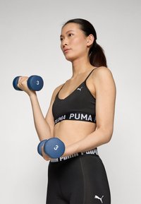 Crni sportski grudnjak s bijelim logotipom "PUMA" i crne tajice visokog struka. Drži plave bučice oznake "3" u neutralnoj postavci.