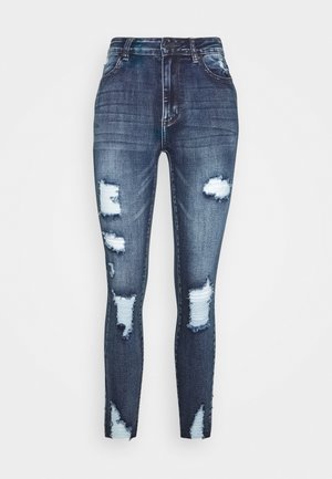 Jean skinny taille haute bleu foncé avec plusieurs patchs blancs usés et trous effilochés sur les jambes avant.