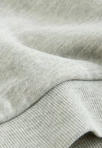 Tessuto di felpa grigio chiaro con una texture liscia, caratterizzato da dettagli a coste e orlo leggermente curvato. Primo piano sui dettagli del materiale.