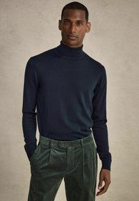 Marinblå tubhalsad tröja i fint stickat tyg, kombinerad med gröna kordbyxor som har knappstängning och ränder.