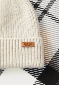 Beanie a coste beige con un'etichetta in pelle marrone con la scritta "Barbour" in rilievo. Sullo sfondo si vede una superficie con un motivo in bianco e nero.