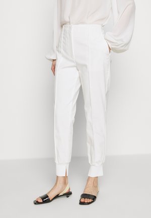 Kobieta ubrana w białe spodnie z wysokim stanem, zwężane, z przednimi zakładkami i podwiniętymi nogawkami, w połączeniu z czarno-beżowymi sandałami na niskim obcasie z odkrytymi palcami.