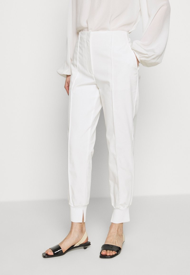 Femme portant un pantalon blanc taille haute à coupe fuselée avec un pli frontal et des ourlets retroussés, assorti de sandales à talons bas ouvertes aux orteils, noires et beiges.