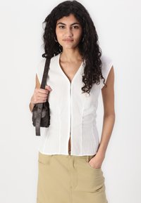 Blouse blanche à manches courtes avec un col en V et une fermeture à boutons sur le devant. Portée avec une jupe kaki, tenant un petit sac noir avec une bandoulière.