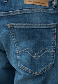 Πίσω τσέπη μπλε τζιν με διπλές ραφές, διακοσμητικό κέντημα και καφέ δερμάτινο ταμπελάκι της μάρκας πάνω από τη μέση.