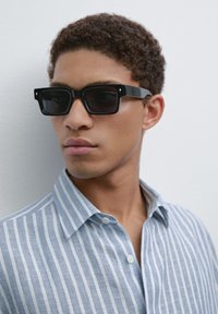 Black rectangular sunglasses with dark lenses, featuring square frames and subtle accent details. Worn with a blue striped button-up shirt.