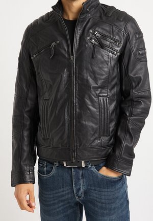 Homme portant une veste en cuir noire zippée avec plusieurs poches zippées et un jean bleu avec ceinture.