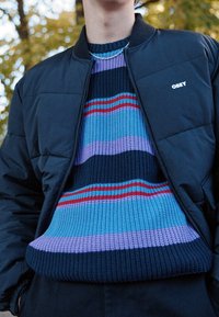 Svart vadderad jacka över en flerfärgad randig stickad tröja med nyanser av blått, lila och rött, med ribbad textur.