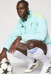 Hele-türkiissinine tõmblukuga jope kollaste triipudega, kombineeritud valgete lühikeste pükstega, põlveni ulatuvate sokkide ja jalgpallikinnitustega, hoides mustrilist jalgpalli.