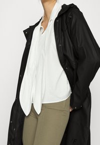 Chaqueta negra resistente al agua con capucha, con botones de presión, llevada sobre una camisa blanca de cuello y pantalones verde oliva. Textura suave.