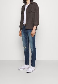 Giacca grigia con bottoni, t-shirt bianca, jeans blu strappati e sneakers bianche. Dettagli notevoli includono due tasche sul petto e una leggera scoloritura.