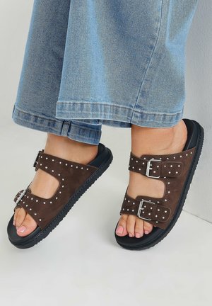 Sandalias de ante marrón con dos correas abatibles, adornadas con tachuelas plateadas y una suela de goma negra. Llevadas con un culotte de mezclilla azul.