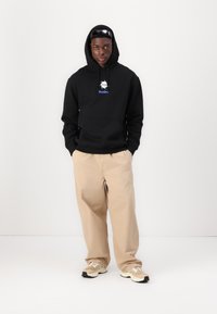 Black hooded sweatshirt with a front pocket and white logo, paired with beige loose-fitting pants and light-colored sneakers.