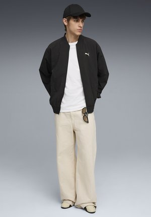 Jonge man met een zwart Puma-jas, wit T-shirt, beige wijde broek, zwarte pet, met een zonnebril in zijn hand, staand tegen een grijze achtergrond.