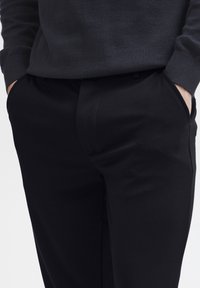 Schwarze Hosen aus einem glatten, strukturierten Stoff mit schmaler Passform, ausgestattet mit Seitentaschen und einem dezenten Knick an der Vorderseite.