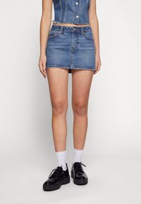 Denim minikjol i ljusblått, med en figurnära design, fransad nederkant och kontrasterande sömmar. I kombination med svarta lackskor och vita strumpor.