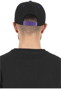 Cappellino nero con un accento viola sulla chiusura a scatto. La texture appare liscia e presenta fori di ventilazione lungo la parte superiore.