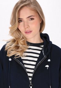 Felpa con zip blu marina con logo a ombrello bianco, indossata sopra una maglietta a righe bianche e nere. Presenta un cappuccio con coulisse e una zip argentata.