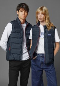 Navy blå puffervästar har en quiltad design med en ståkrage, kontrasterande röda dragkedjor och logoplagg på bröstet. Bärs över vita t-shirts.