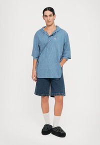 Camisa a cuadros azul con capucha, combinada con pantalones cortos de mezclilla y zapatos negros sin cordones. Características incluyen un corte holgado y una correa cruzada.