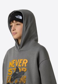 Hall halliga kapuutsiga pusa, millel on oranž graafika tekstiga "NEVER STOP EXPLORING", ja must soonikust nokamüts kaubamärgi logoga.