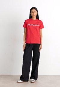 Červené bavlnené tričko s bielym logom "Calvin Klein Jeans", skombinované s voľnými čiernymi nohavicami a striebornými teniskami. Hladká textúra a uvoľnený strih.
