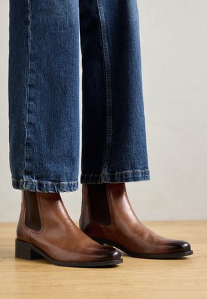 Botas de tornozelo de couro castanho com painéis laterais elásticos, bico quadrado e salto em bloco baixo, usadas com jeans de ganga azul. Chão em madeira abaixo.