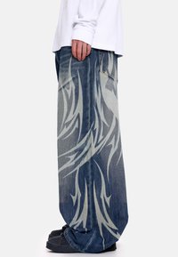 Τζιν με ευρύ πόδι, σε μπλε ντενίμ με αντίθετο ασημί σχέδιο φλόγας. Ξεθωριασμένη υφή και μία τσέπη στο πίσω μέρος.