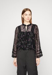 Blusa nera trasparente con stampa floreale, dettagli in pizzo e scollatura con lacci. Caratterizzata da maniche lunghe e una vestibilità morbida, abbinata a pantaloni a righe.