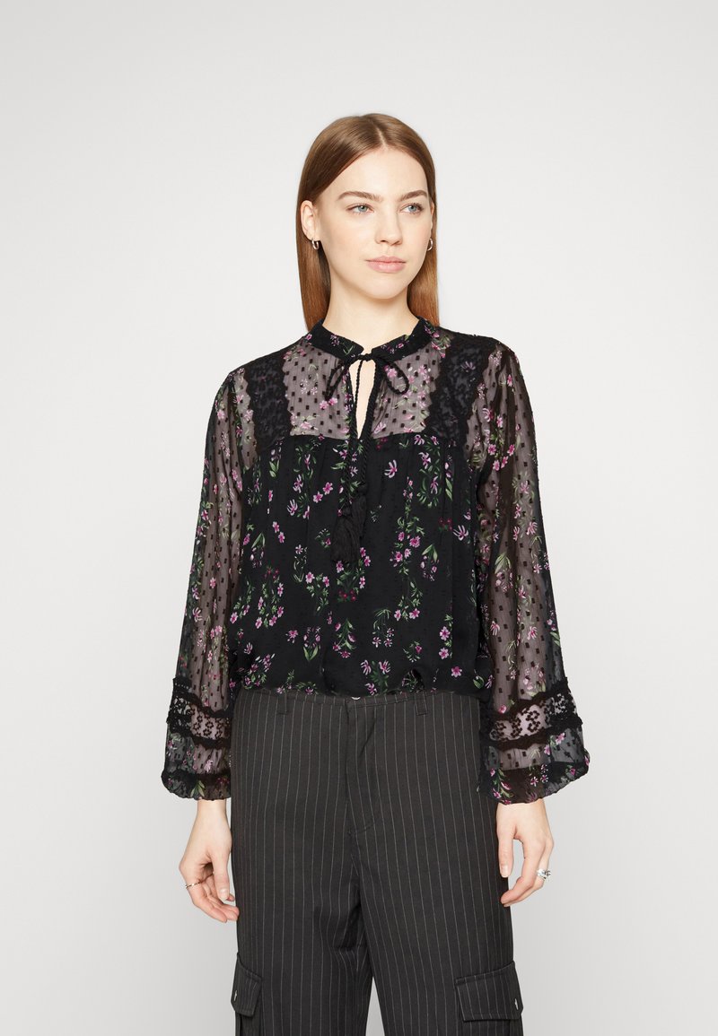 Blusa nera trasparente con stampa floreale, dettagli in pizzo e scollatura con lacci. Caratterizzata da maniche lunghe e una vestibilità morbida, abbinata a pantaloni a righe.