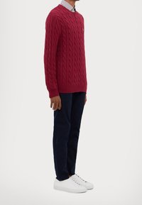 Röd kabelstickad tröja med rund halsringning, ribbade muddar och nederkant, bärs över en randig skjorta med krage, tillsammans med mörkblå byxor och vita sneakers.