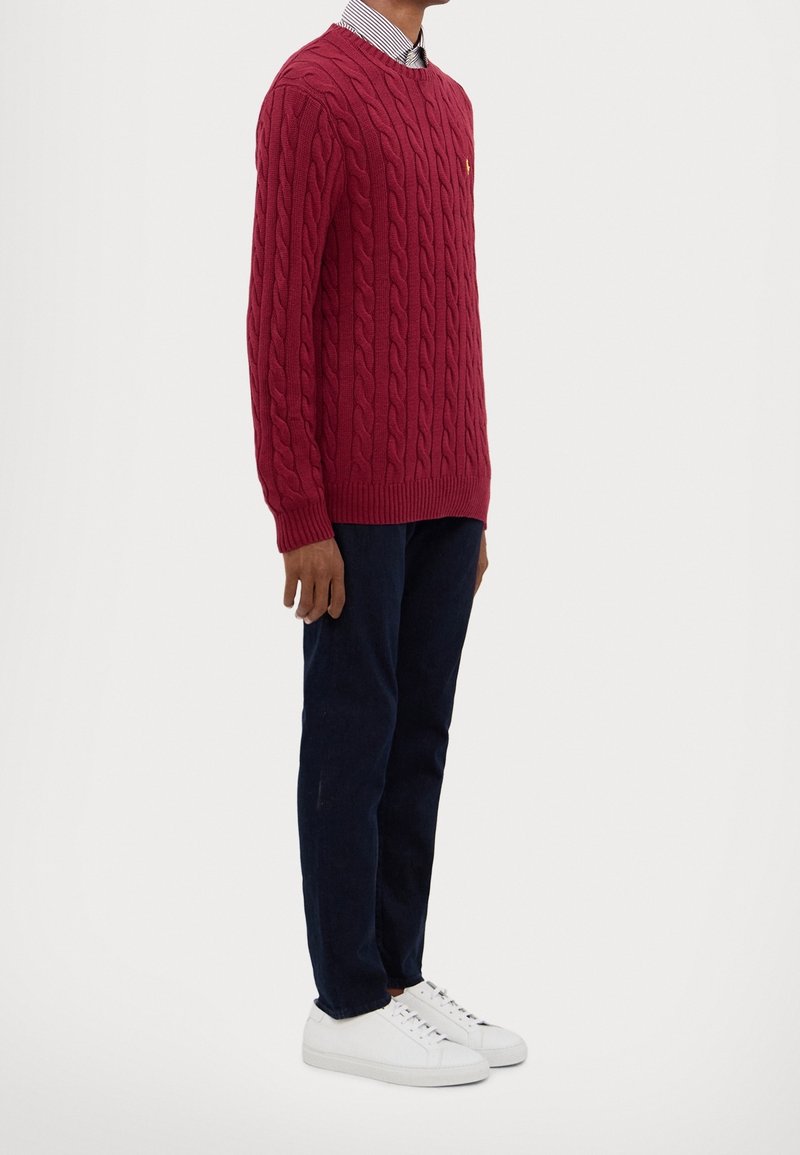 Röd kabelstickad tröja med rund halsringning, ribbade muddar och nederkant, bärs över en randig skjorta med krage, tillsammans med mörkblå byxor och vita sneakers.