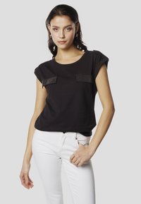 Must lühikeste varrukatega T-särk, millel on kaks tekstuuriga taskut metallilise detailiga. Sobitatud valgete kitsaste teksapükstega kontrasti loomiseks.