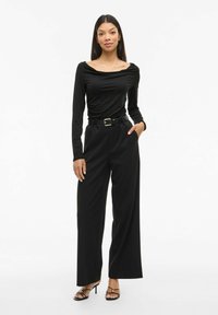 Zwarte lange mouw top met een coltrui, gecombineerd met hoge zwarte wijde broek en zwarte sandalen met bandjes met gouden accenten.