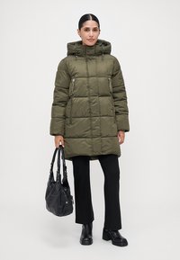 Mujer vestida con un abrigo acolchado con capucha verde oliva, pantalones negros y botas negras, sosteniendo un bolso negro frente a un fondo liso.