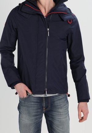 Mężczyzna ubrany w granatową kurtkę zapinaną na zamek z czerwonymi akcentami i wyblakłe niebieskie dżinsy, z ręką w kieszeni, stoi na tle jednolitego tła.