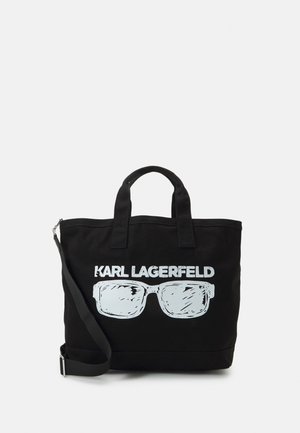 Czarna płócienna torba na ramię z uchwytami i regulowanym paskiem. Na przodzie duży biały nadruk okularów przeciwsłonecznych oraz napis „KARL LAGERFELD” poniżej.