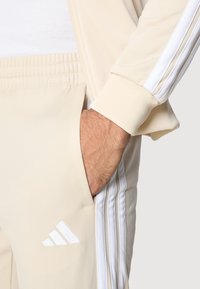 Χέρι στην τσέπη μπεζ φόρμας Adidas με λευκές ρίγες και λογότυπο, συνδυασμένη με αντίστοιχο μπεζ μπουφάν με λευκές ρίγες.