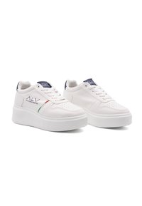 Valkeat nauhalla varustetut tennarit, joissa on litteä ja paksu pohja. Kenkien sivuissa on reikiä, ja niissä on tummansinihärppä sekä vihreät ja punaiset raidat.