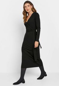 Schwarzes Wickelkleid mit langen Ärmeln, V-Ausschnitt und Raffung. Weicher Stoff, Midi-Länge, mit gebundenem Taillenbereich, kombiniert mit schwarzen Ballerinas und Strumpfhosen.