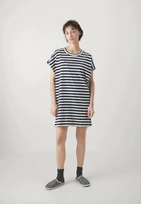 Kobieta z krótkimi włosami nosi luźną sukienkę w czarno-białe paski z nadrukiem, czarne skarpetki i koszule w kratę, stojąc na jednolitym tle.