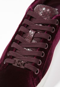 Baskets en velours bordeaux avec lacets tissés, languette à texture de peau de serpent et breloque logo MK métallique sur une semelle blanche.