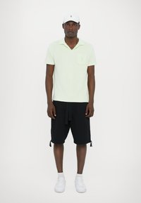Lichtgroene polo met een zak, gecombineerd met zwarte shorts met een trekkoord. Witte sneakers en een witte pet maken de outfit compleet.