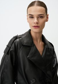 Cappotto trench in pelle nera con chiusura doppiopetto, grandi revers e spalline. Texture liscia con minimalismo nei dettagli.