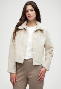 Lažni krzneni skraćeni kaput u krem boji, s ovratnikom, prednjim gumbom za zatvaranje i dugim rukavima. Tkanina ima mekan, teksturiran izgled.