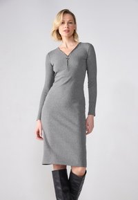 Robe en tricot côtelé grise avec des manches longues, un décolleté en V et une fermeture éclair à l'avant, tombant jusqu'au mollet, assortie à des bottes noires hautes.